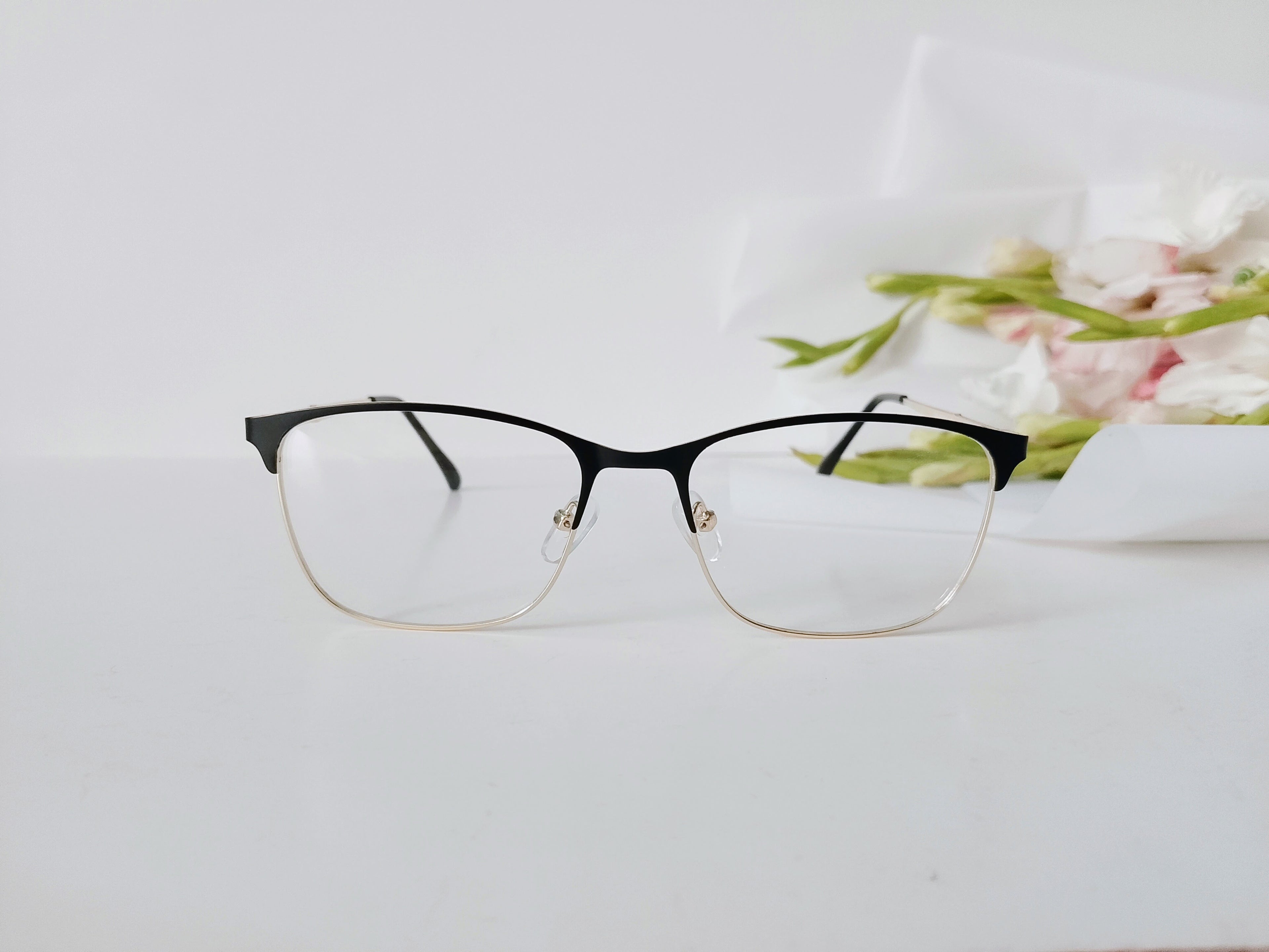 Black and gold semi-rimless eyeglasses with clear lenses on white surface, floral background