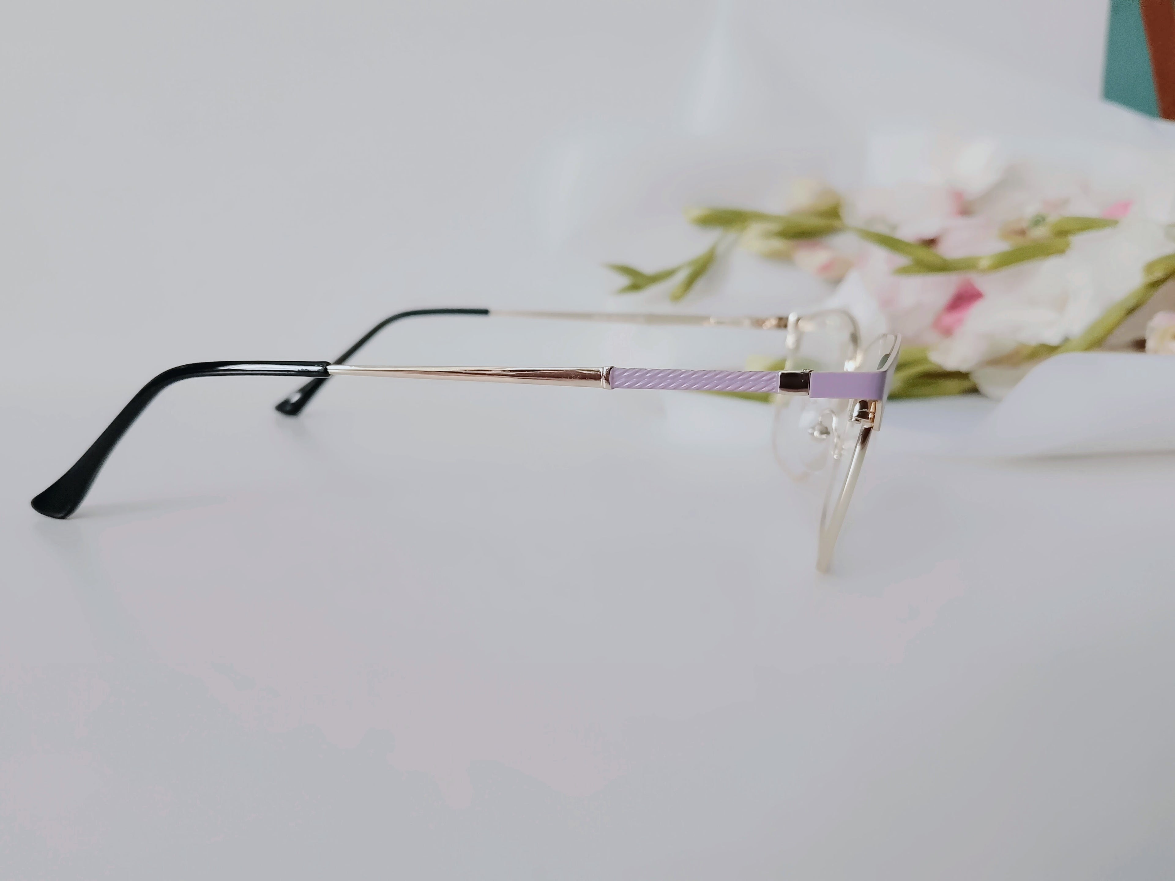 Elegant rimless glasses with lavender temple accents, sleek black arms, and gold detailing on white surface with floral backdrop