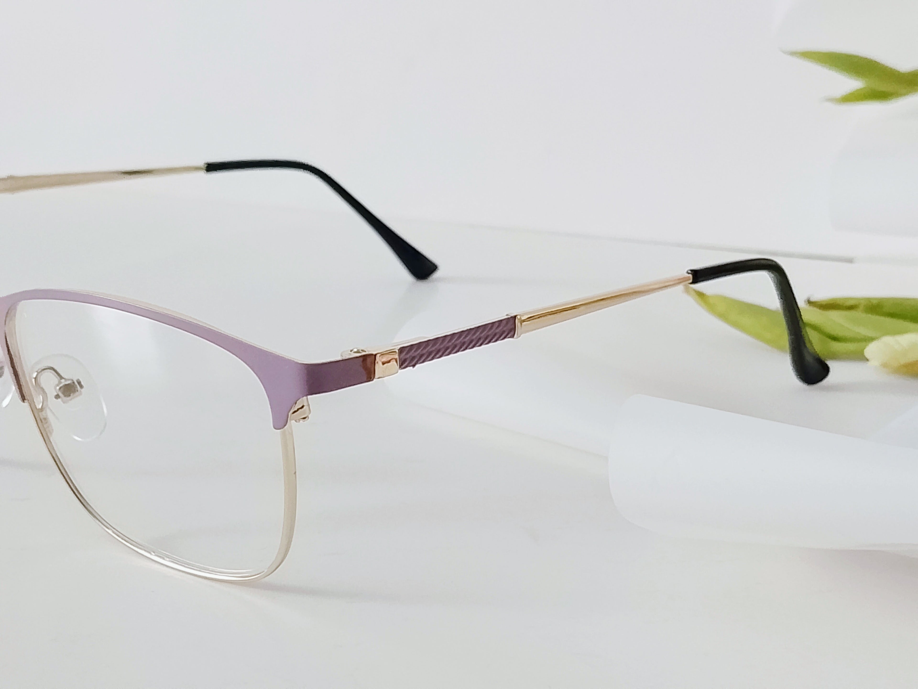 Purple and gold rimless eyeglasses with clear lenses, sleek modern design on white surface with green leaf accent
