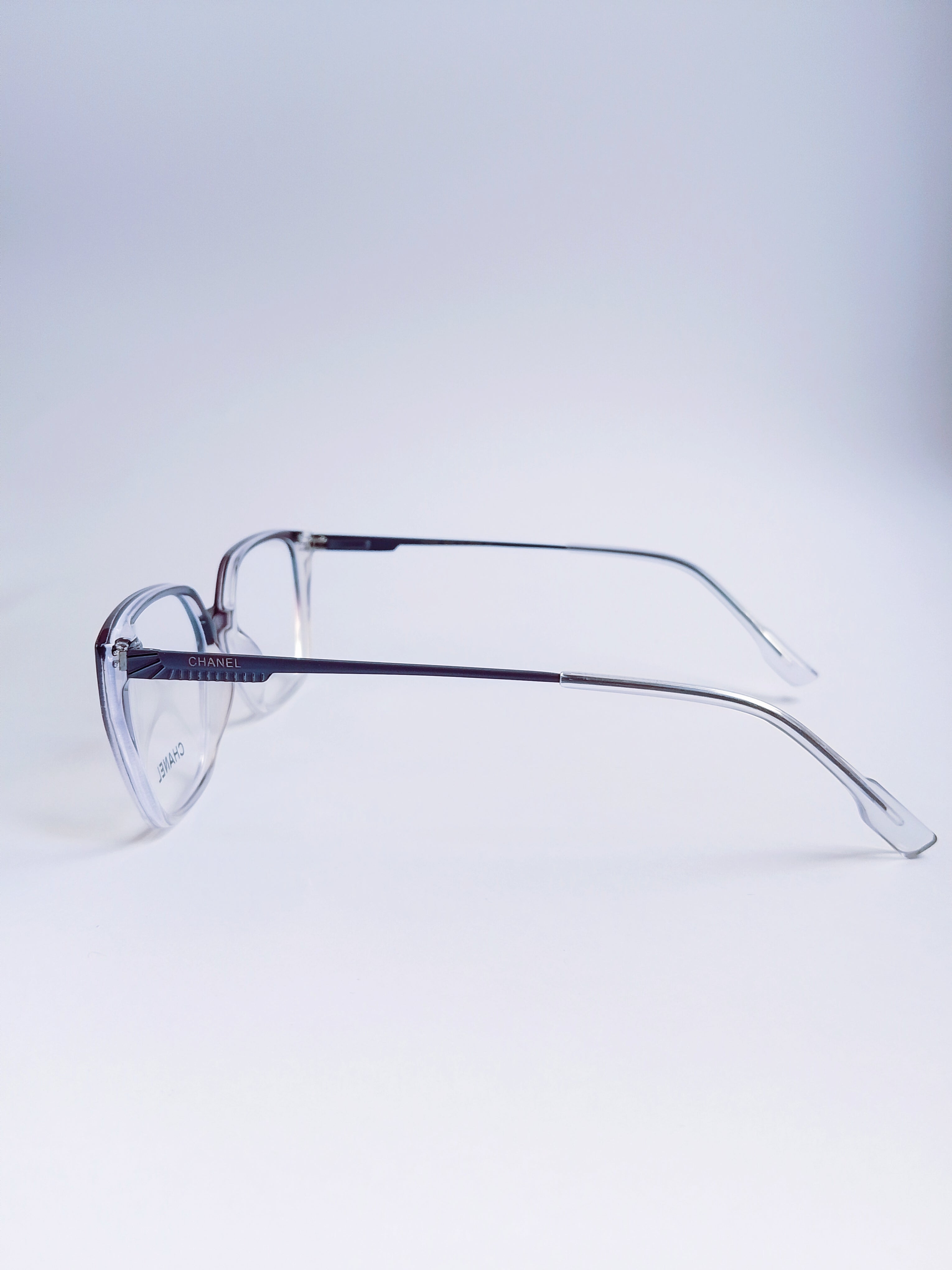 A pair of clear acetate eyeglass frames with dark metal temples, shown from a side profile on a light gray studio background.