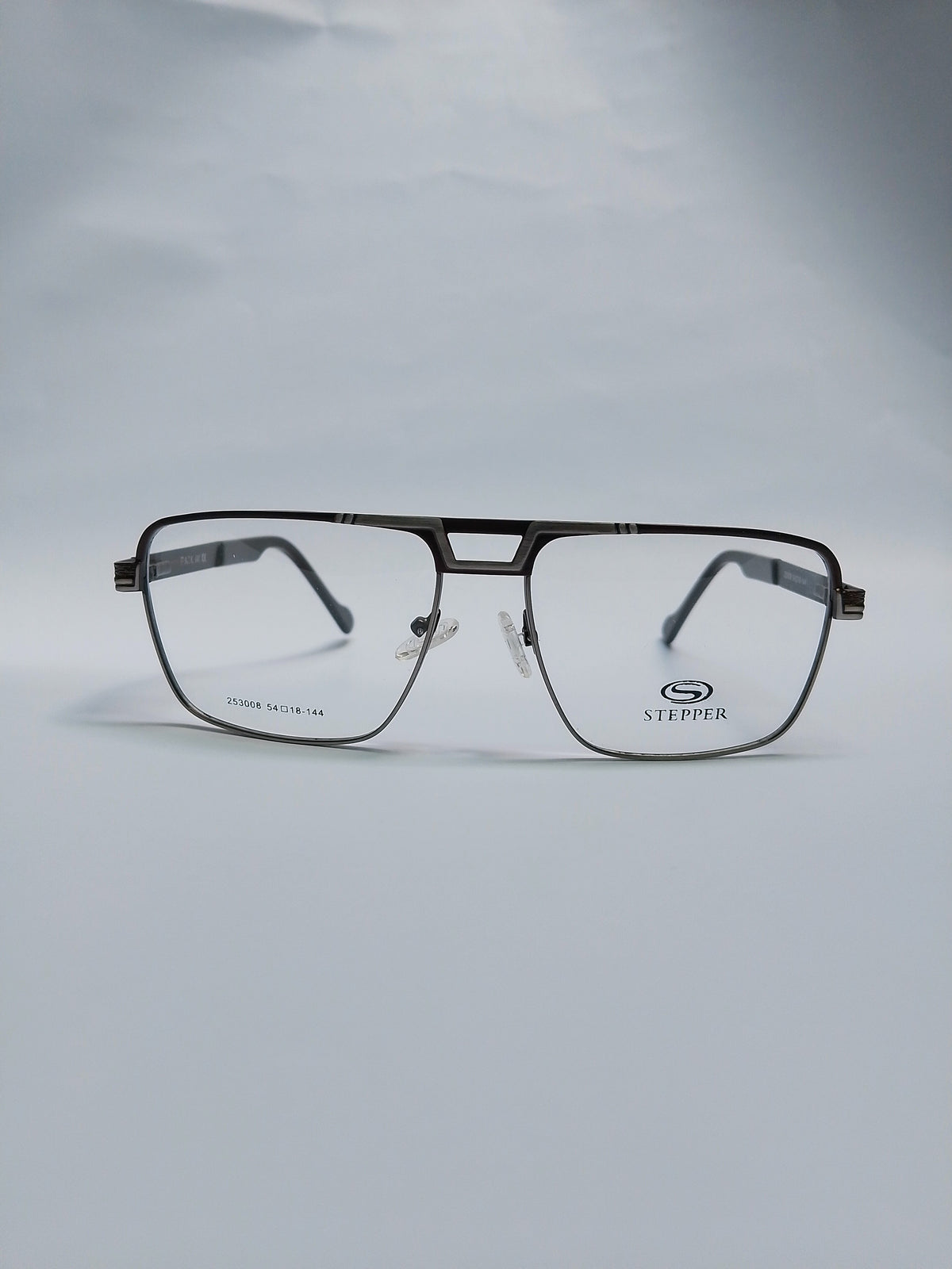 A pair of aviator-style eyeglasses with a thin black metal frame and clear lenses, resting on a light gray surface against a matching gray background. The brand name 'STEPPER' is visible on the lower right lens.