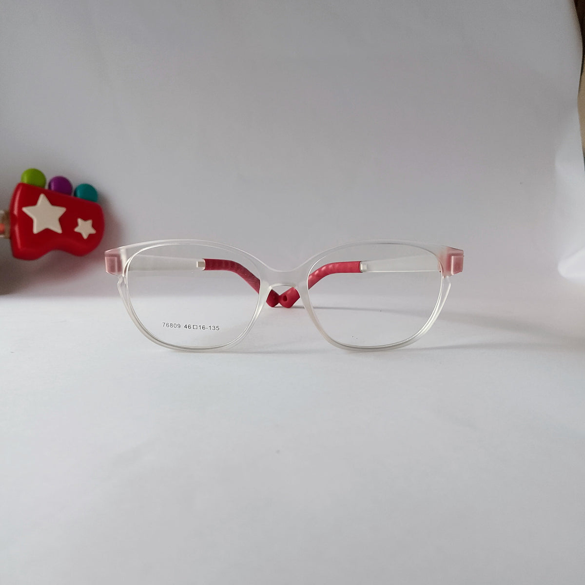 Clear acetate eyeglass frames with red inner temples and nose bridge, shown on a white background with a small red toy prop on the left.