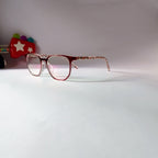 A pair of round eyeglasses with a translucent pink and red acetate frame and patterned temples, resting on a white surface against a light background.