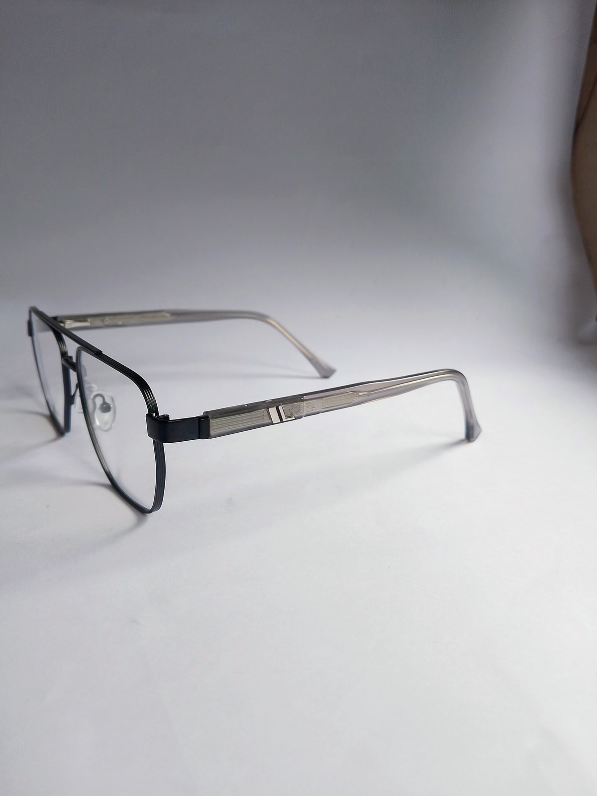 A pair of square aviator-style eyeglasses with a black front frame and silver metal temples, resting on a white surface against a gray background.