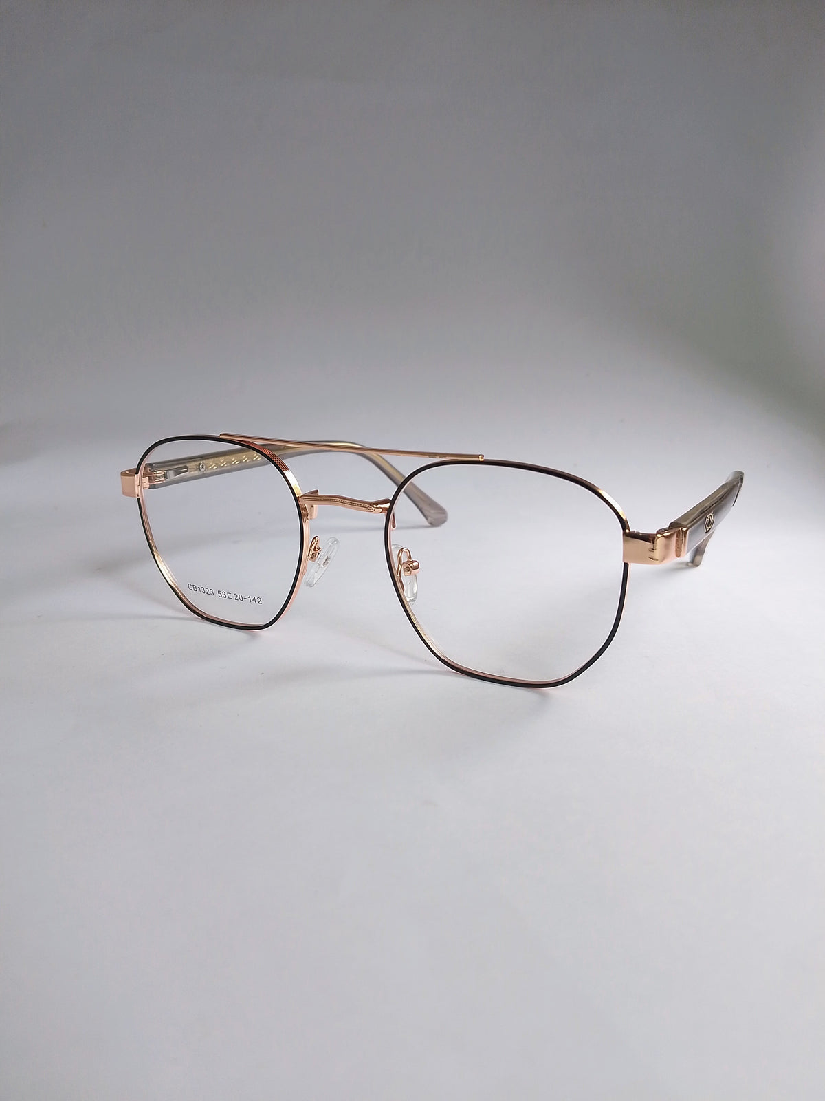 A pair of aviator-style eyeglasses with a thin rose gold metal frame and clear lenses, resting on a light gray surface against a neutral gray background.
