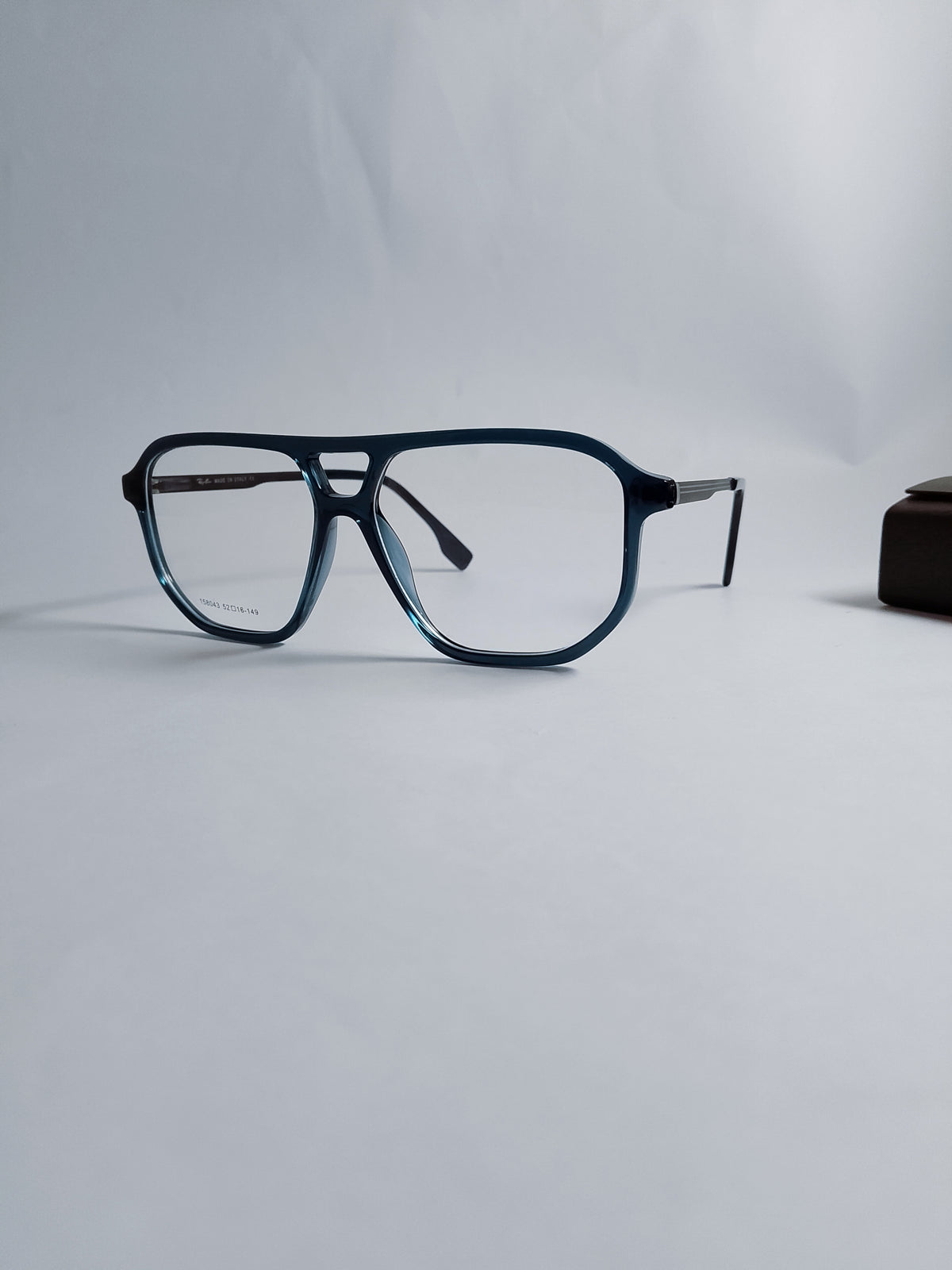 A pair of rectangular eyeglasses with a dark blue acetate frame and clear lenses, displayed on a light gray background with a small dark accessory partially visible on the right.