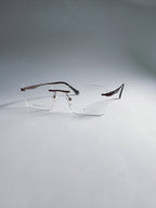 A pair of rimless eyeglasses with a thin metal frame and clear lenses, resting on a light gray surface against a matching gray background.