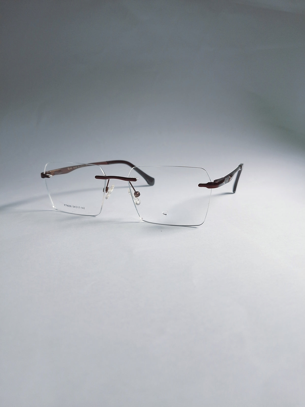 A pair of rimless eyeglasses with a thin metal frame and clear lenses, resting on a light gray surface against a matching gray background.