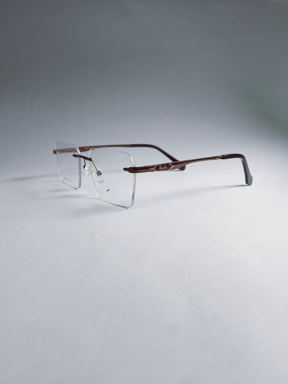 A pair of rimless eyeglasses with a dark brown metal frame and clear lenses, displayed on a light gray studio background.