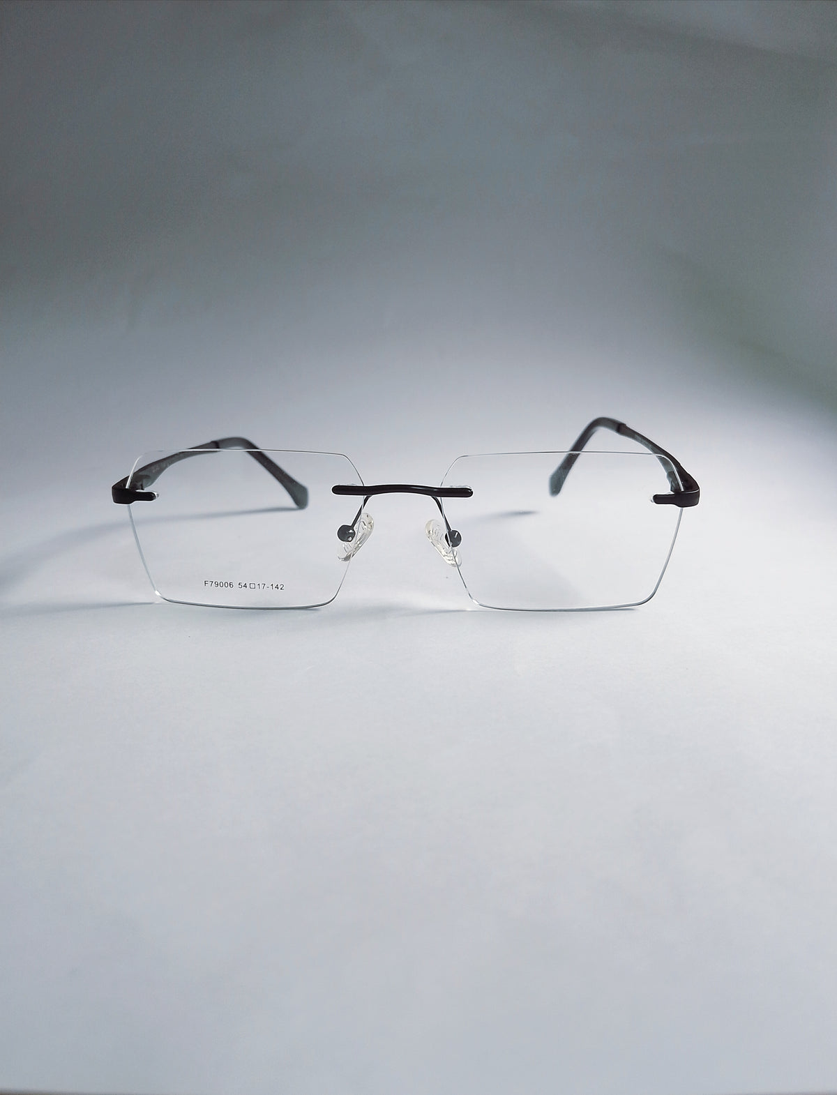 A pair of rimless eyeglasses with a thin black metal frame and clear lenses, displayed on a light gray background.