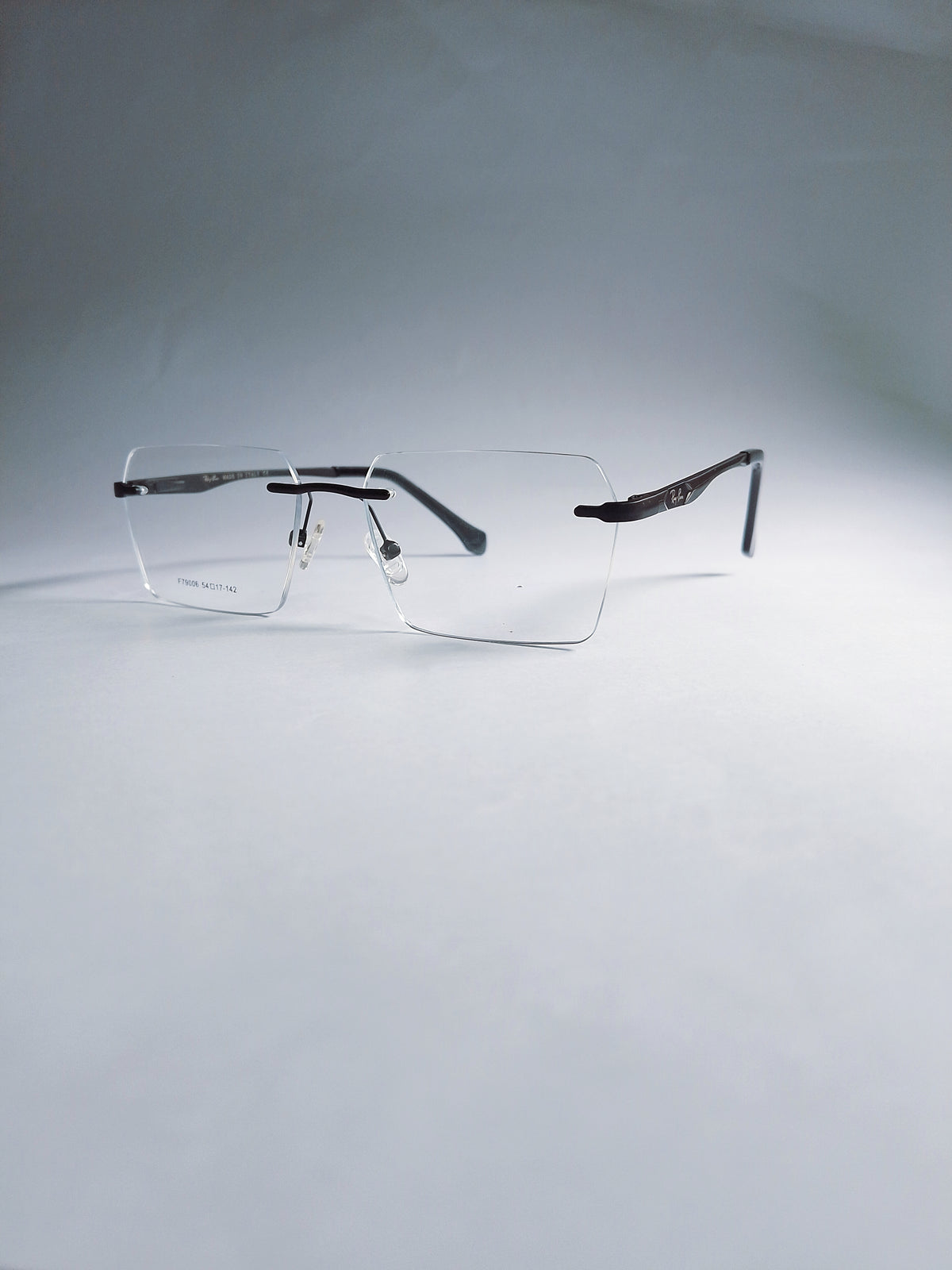 A pair of rimless square eyeglasses with clear lenses and dark metal temples, displayed on a light gray gradient background.