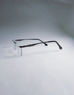 A pair of rimless eyeglasses with dark brown arms and clear lenses, displayed on a clear stand against a light gray background.