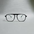 Black aviator-style eyeglasses with clear lenses and green temple tips, resting on a light gray surface.